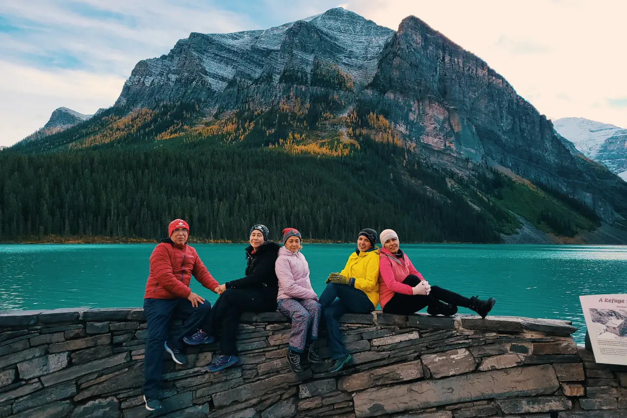 Five individuals sitting on a stone wall with a mountainous backdrop and turquoise lake.