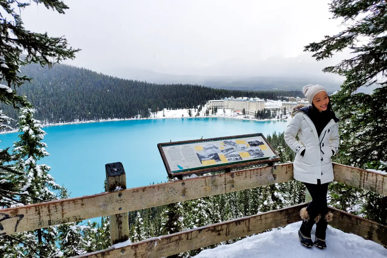 A person in winter clothing stands by an information sign overlooking a turquoise lake and a large hotel.