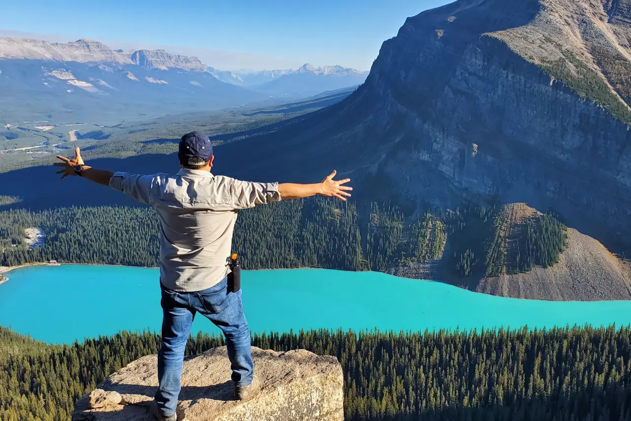 Man with arms spread overlooking a turquoise lake and mountains.