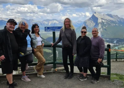 A group of six people poses at an overlook sign indicating 2085m elevation, with stunning mountains and valleys in the background.